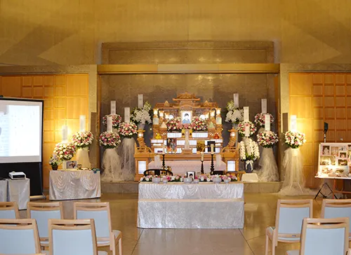 葬儀事例「おばあちゃんへの恩返し」のギャラリー画像3枚目 正面に白木祭壇と多数の供花、上手には大型スクリーンが設置